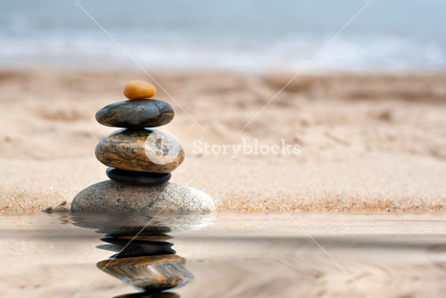 A Pile Of Round Smooth Zen Like Stones Stacked In The Sand At The Beach With A Mirror Reflection From A Pool Of Water.